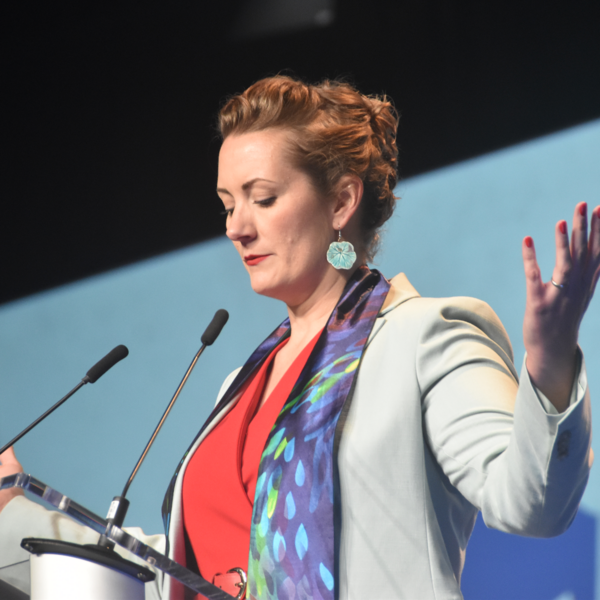 a pastor stands behind a podium with her arms stretched out and head bowed
